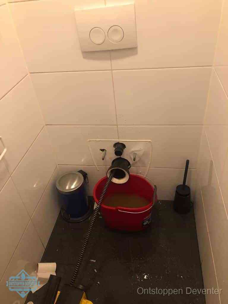 Een ontstopte wand-wc met verwijderde toiletpot waar een ontstoppingsveer in de wanduitgang verdwijnt, het achterhalen waar de verstopping zit in Deventer