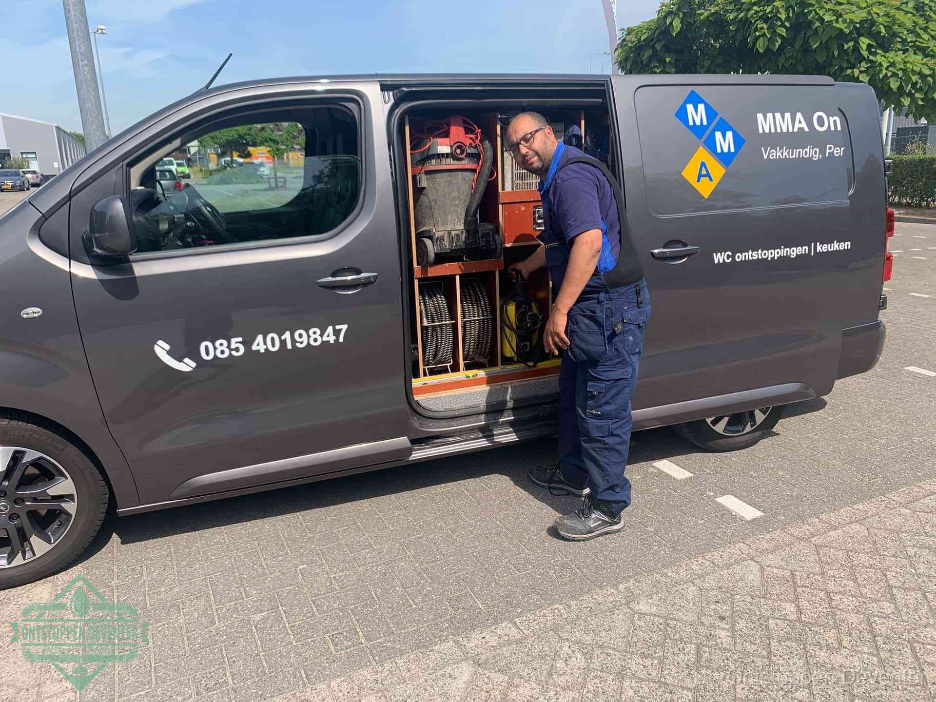 Een monteur bij de open schuifdeur van zijn bedrijfsbus op een parkeerplaats, klaar om de oorzaak van terugkerende afvoerverstoppingen aan te pakken in Deventer